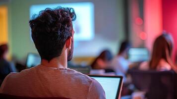 Young man with curly hair is focused on laptop during a seminar in a modern classroom, with blurred audience and colorful lighting creating an engaging atmosphere photo