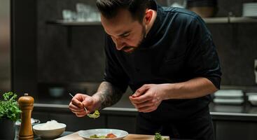 Male chef in black uniform meticulously plating gourmet dish in modern kitchen, showcasing culinary skills and attention to detail in a professional environment photo