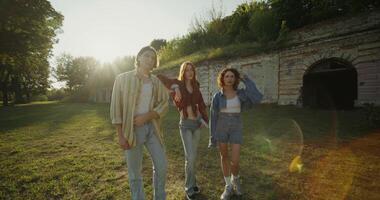 Three young friends look into the camera with a calm look standing near the ruins of a historic building. The fortress walls are overgrown with green vegetation. A sunny summer day photo