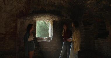 Three friends smile and talk as they walk around a room in a historic abandoned brick building. Sunlight photo