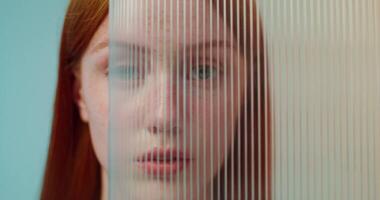 A red-haired woman with freckles is smiling at the camera while standing on a studio background. She hides her face behind plexiglass photo