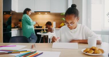 A girl draws with a stylus on a tablet while sitting at the kitchen table at home. Her family in the background is out of focus - sitting photo