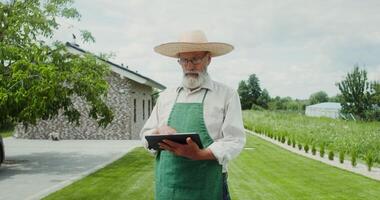 An elderly man with a gray beard, dressed in an apron and hat, smiles while looking at the camera standing in the backyard with a tablet in his hands photo