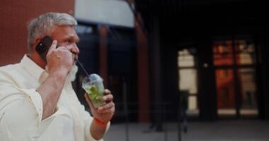 A gray-haired man with a mustache and beard talking on a mobile phone and drinking lemonade while walking along a city street photo
