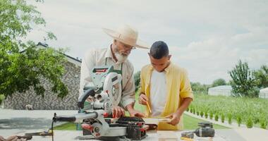 An elderly man teaches his grandson how to use a miter saw while standing in the backyard of his house - standing photo