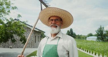 An elderly man with a gray beard, dressed in an apron and hat, smiles while looking at the camera standing in the backyard with a rake in his hand - smiles photo