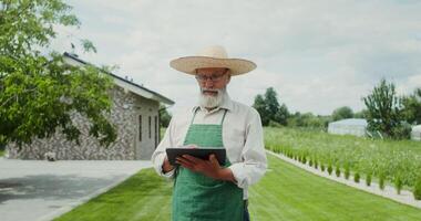An elderly man with a gray beard, dressed in an apron and hat, smiles while looking at the camera standing in the backyard with a tablet in his hands - smiles photo