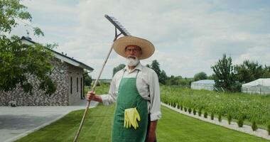 An elderly man with a gray beard, dressed in an apron and hat, smiles while looking at the camera standing in the backyard with a rake in his hand photo