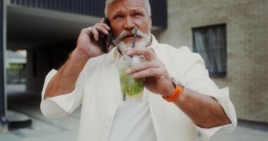 A gray-haired man with a mustache and beard talking on a mobile phone and drinking lemonade while walking along a city street - drinking photo