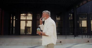 A gray-haired man with a mustache and beard talking on a mobile phone and drinking lemonade while walking along a city street - mobile photo
