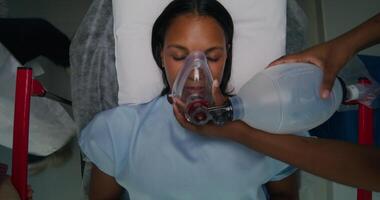 The patient is lying on a gurney with her eyes closed. The nurse gives her air through an oxygen mask. View from above. Emergency photo