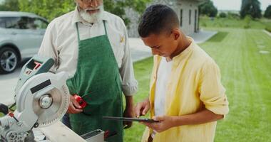 Grandfather and grandson are talking and using a tablet while standing in the backyard near a miter saw photo