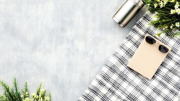 A flat lay displays a thermos, notebook, sunglasses, and blooming flowers arranged on a picnic blanket, suggesting a leisurely outing photo