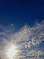 Celestial Canvas - A clear blue sky adorned with fluffy white clouds, where sunshine radiates from behind and casts a warm, inviting glow across the horizon. photo