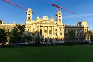 Front view of Museum of Ethnography in Budapest with neoclassical architecture, wide green lawn and construction cranes under clear blue sky photo