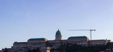 Wide view of Buda Castle in Budapest with green dome and surrounding wings under clear blue sky, with construction crane visible in the background photo