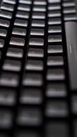 A close-up of a sleek black keyboard with square keys, resting on a dark, textured surface. The keyboard's sharp angles and minimalist design create photo