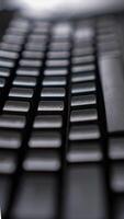 A close-up of a sleek black keyboard with square keys, resting on a dark, textured surface. The keyboard's sharp angles and minimalist design create a modern photo