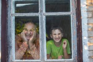 Grandfather and grandson look out of the window of a village house and grimace. Father's Day. photo