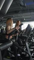 Beautiful young women are chatting while exercising on elliptical trainers or cross-trainers in gym. They move slowly and gracefully. video
