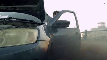 Young driver of car panics and runs out to look under the hood, from where black smoke is billowing. This indicates possible engine problem. Slow motion. video