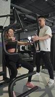 Charming young athlete is performing chest exercises in gym, under guidance of her personal trainer. Trainer has youthful Asian appearance and is monitoring effectiveness of exercises. video