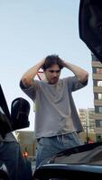 Young man stands on city road next to open hood of car, with his head in his hands. He is worried or disappointed because of mechanical malfunction that occurred after accident. video