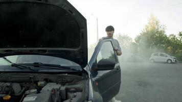 Driver gets out of car, surrounded by smoke, and tries to figure out what caused breakdown. They look under open hood, in middle of city parking lot. video