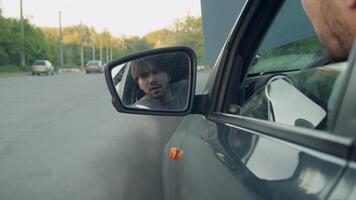 Man leans out of car window on driver's side. His face is reflected in side mirror when black smoke starts pouring out of car. He runs to put out fire with fire extinguisher in his hand. video