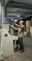 Two men in protective helmets and glasses are talking standing near the machine control panel at a paper mill photo
