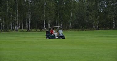 Side view of a golf cart driving through a golf course with two men and equipment. Well-groomed golf course photo