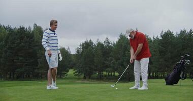 Father and son play golf together, talking and laughing. A young handsome man of European appearance looks as an elderly gray-haired man with a beard is hitting ball with a golf club. photo