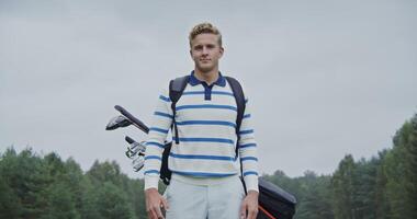 young handsome blond man of European appearance, dressed in sportswear with golf equipment in a bag behind his back, stands on the golf course and looks at the camera smiling photo