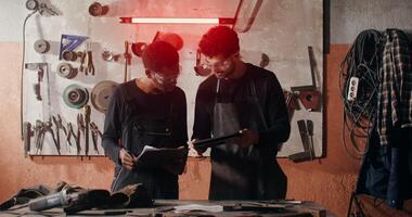 Two blacksmiths of different nationalities are thinking about a sketch of a future product, working together and using a tablet to search for ideas, in workshop full of tools. in 4k, red komodo photo