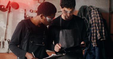 Two blacksmiths of different nationalities are thinking about a sketch of a future product, working together and using a tablet to search for ideas, in workshop full of tools photo