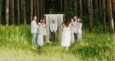 The bride walks to the altar passing by the applauding guests. The groom is waiting for her standing near the priest. Wedding in the forest photo