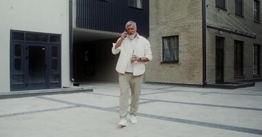A gray-haired man with a mustache and beard talking on a mobile phone and drinking lemonade while walking along a city street - lemonade photo