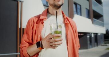 A stylish young man enjoys drinking mojito from a disposable cup with a straw while standing on a city street photo