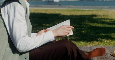 Young beautiful woman is reading a book sitting on a blanket on the riverbank near the red suspension bridge photo