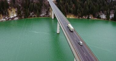 View of a concrete road bridge with a two-lane asphalt road with cars passing on it. drone footage of a long bridge across an emerald green river to hills covered in snowy forest. photo