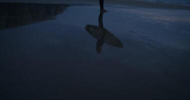 Surfer with a board in his hands stands on the seashore at dusk, reflected in the water on the shore photo