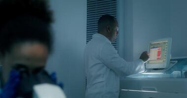 A man in a lab coat sets up a blood splitting machine. A woman uses a microscope sitting at a table. photo