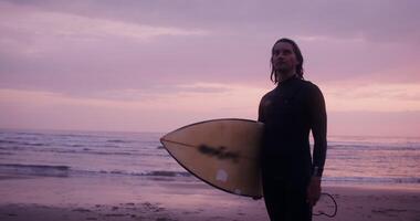 A man with wet hair and a surfboard in his hands, stands on the seashore in the red light of the setting sun photo