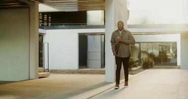 A young stylishly African-American man with a laptop case in his hand drinks coffee from a disposable cup while walking around the courtyard of a modern building. in 4k, red komodo photo
