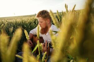 Country musician is,, with guitar, on the ground. Man in white shirt is on the agricultural field photo