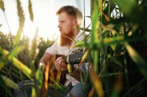 Country musician, is with guitar on the ground. Man in white shirt is on the agricultural field photo