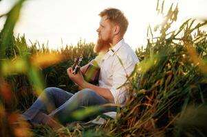 Country musician is with guitar ,on, the ,ground. Man in white shirt is on the agricultural field photo