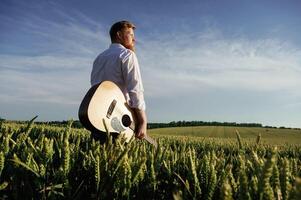 Musician is with acoustic guitar. Man, in ,white shirt is on the agricultural field photo
