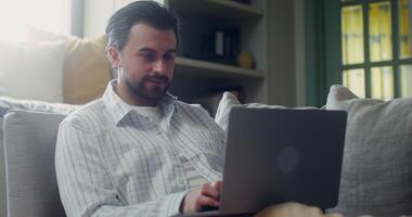Stylish man in casual clothes smiles typing on a laptop while sitting on a sofa indoors photo