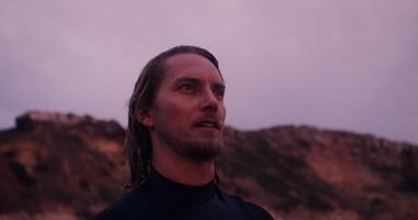 Close-up of face of a young man in a diving suit, with wet hair, standing near a rock in the open air at dusk photo